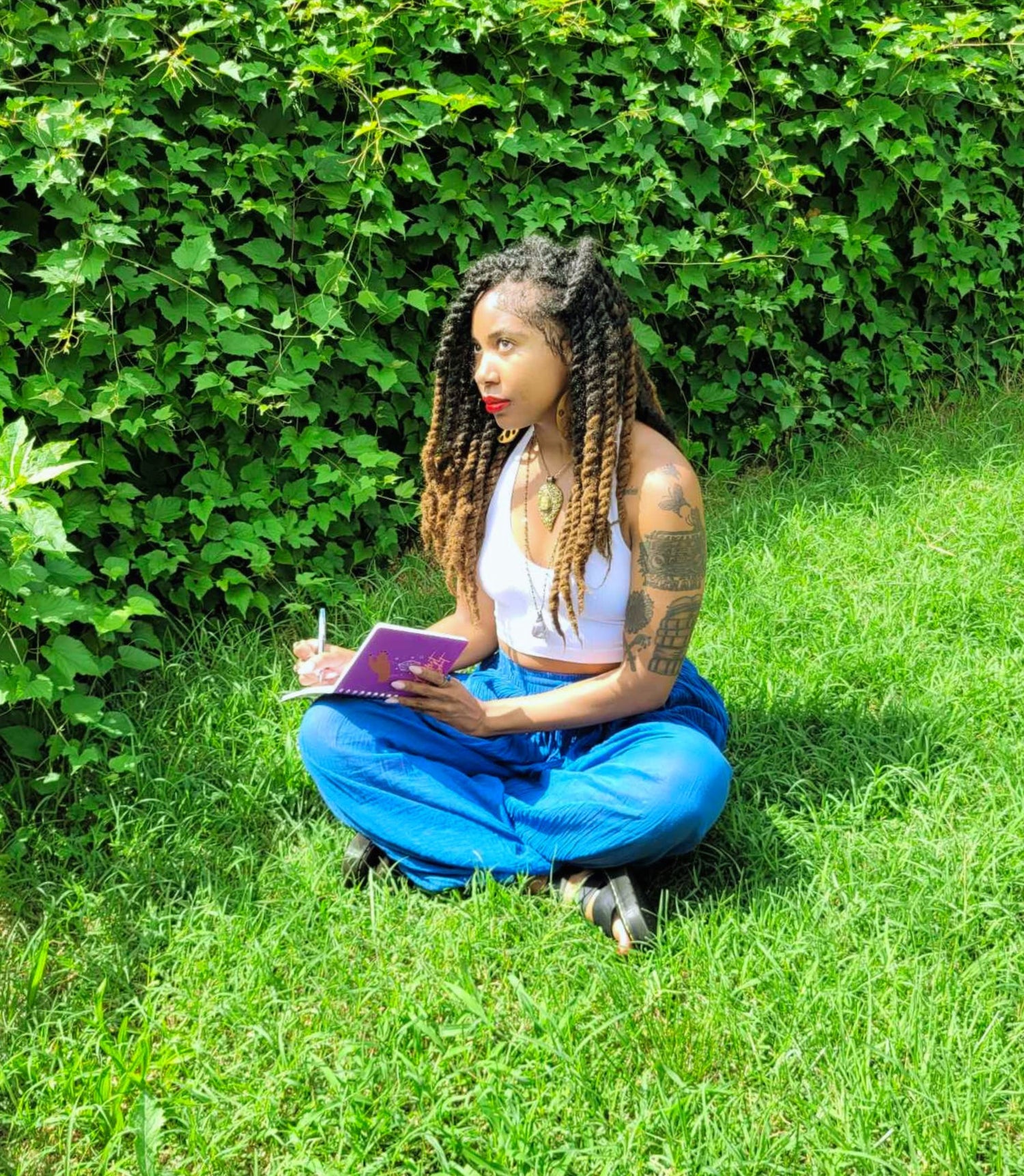Woman sitting on grass with a notebook and pen, surrounded by greenery