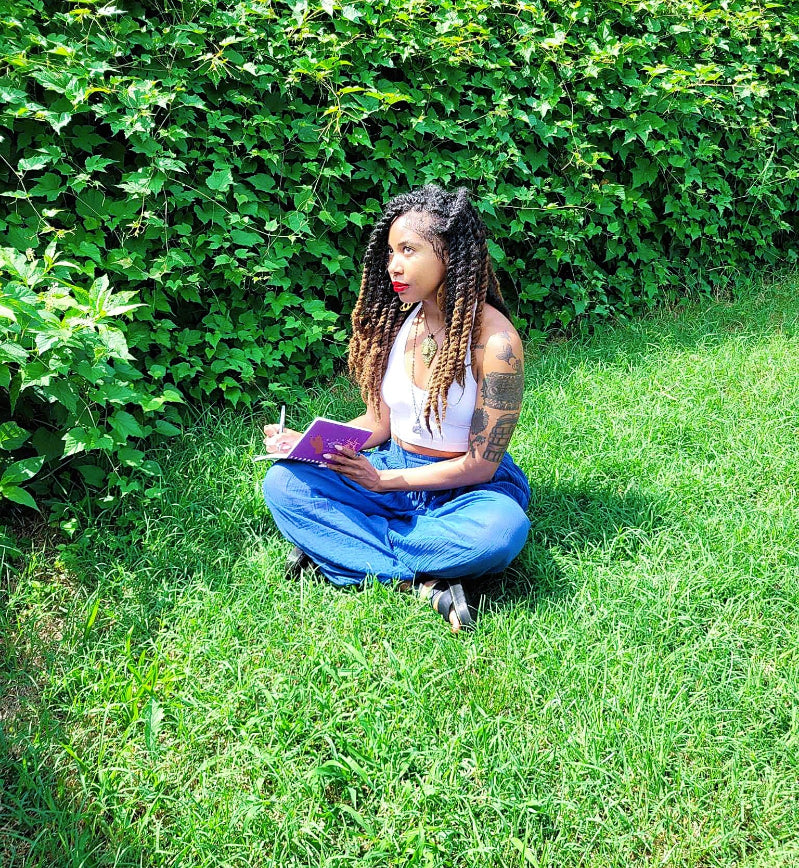 Woman sitting on grass holding a purple notebook with greenery in the background
