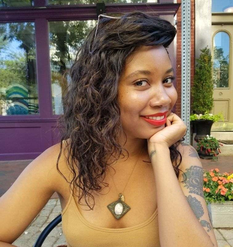 Woman with tattoos and a necklace sitting outdoors, smiling.