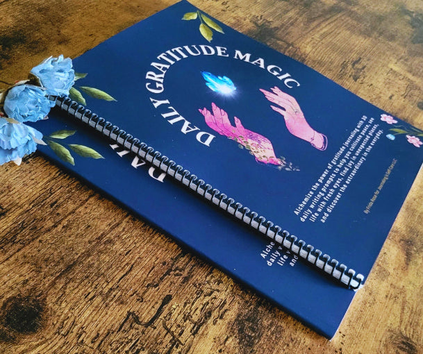 Blue spiral-bound book titled 'Gratitude Magic' with floral decorations on a wooden surface