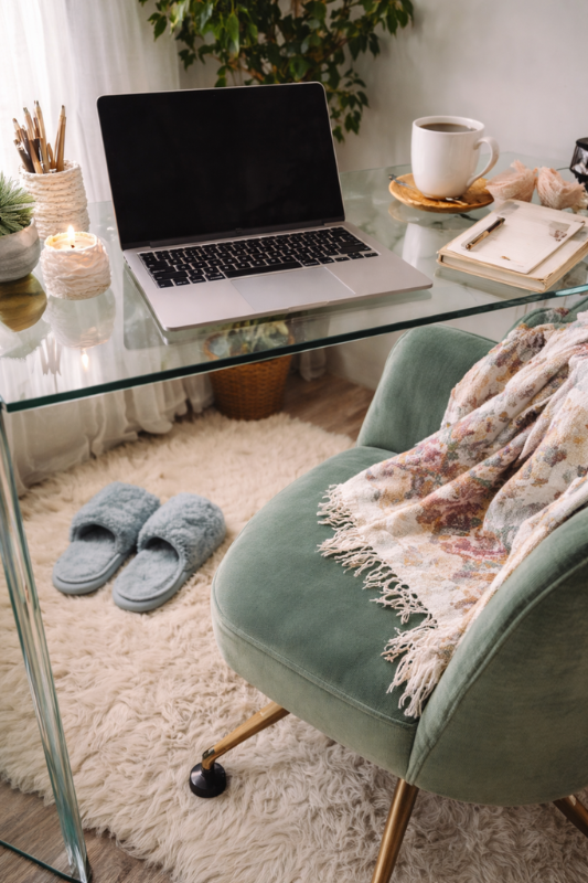 Home office setup with a laptop on a glass desk, chair with a blanket, and slippers.