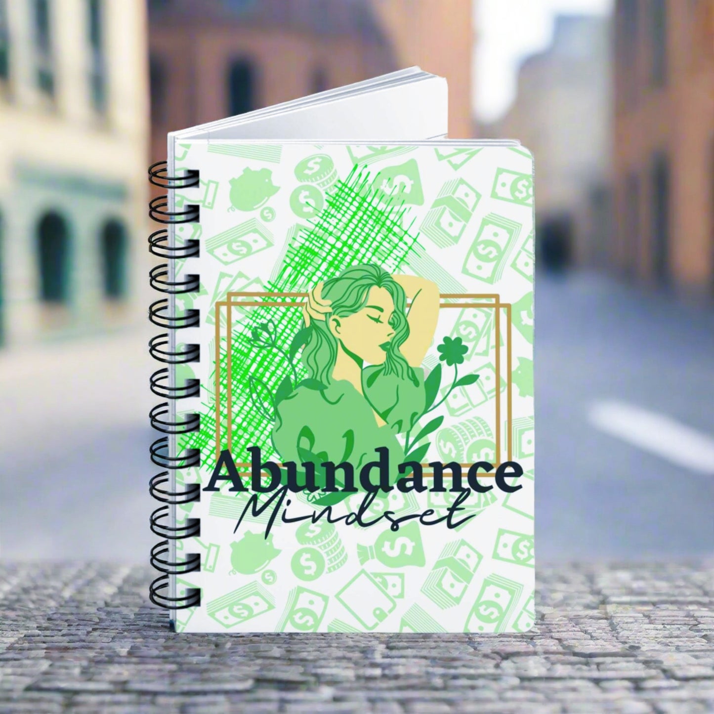 A spiral notebook with the words Abundance Mindset Journal with Journal Prompts on it.