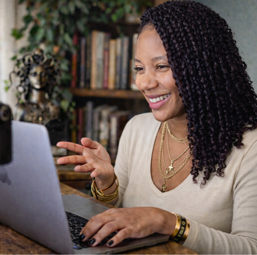 Woman sitting at a desk with a laptop, smiling and interacting with a camera on a tripod.