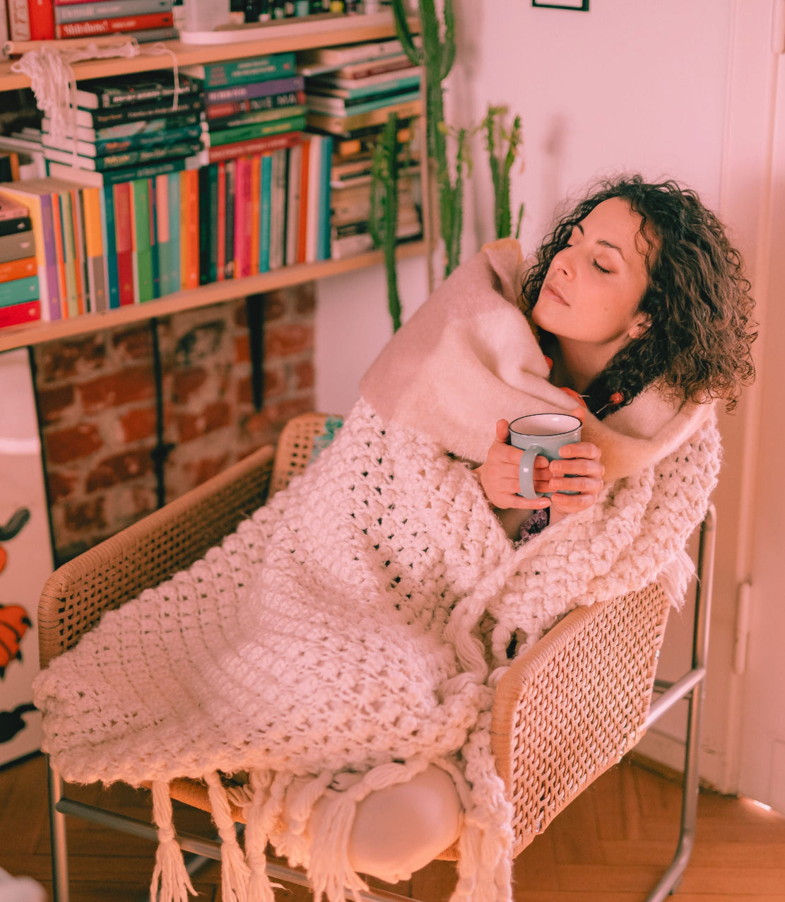 A self-care moment for a woman wrapped in a blanket holding a mug in a cozy room with books and plants.