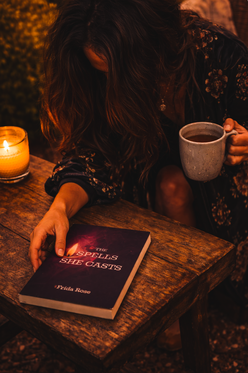 Person reading a book titled 'The Spells She Casts' with a candle and mug on a wooden table.