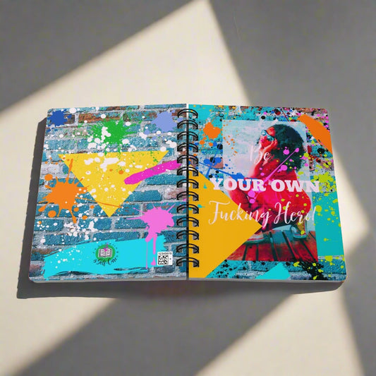 A "Be Your Own Fucking Hero" Spiral Bound Journal for Black Women with colorful splatters on it, perfect for self-reflection and new beginnings.