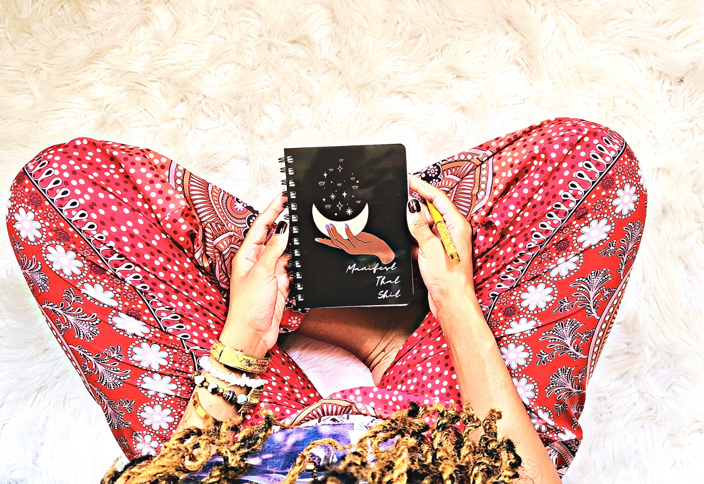 A woman is sitting on a rug with a "Manifest That Shit" Manifesting Journal to Access Your Inner Power on her lap.